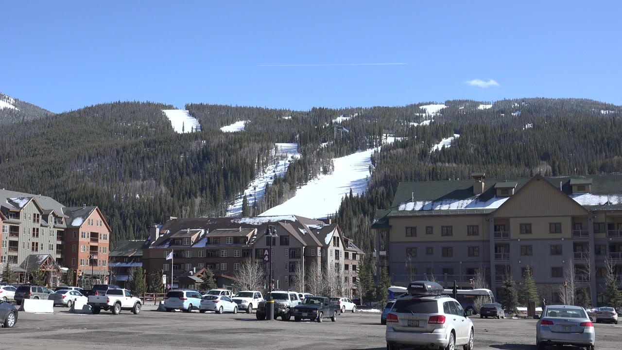 Wide view of cars, lodge buildings, and snowy ski runs on a mountain.