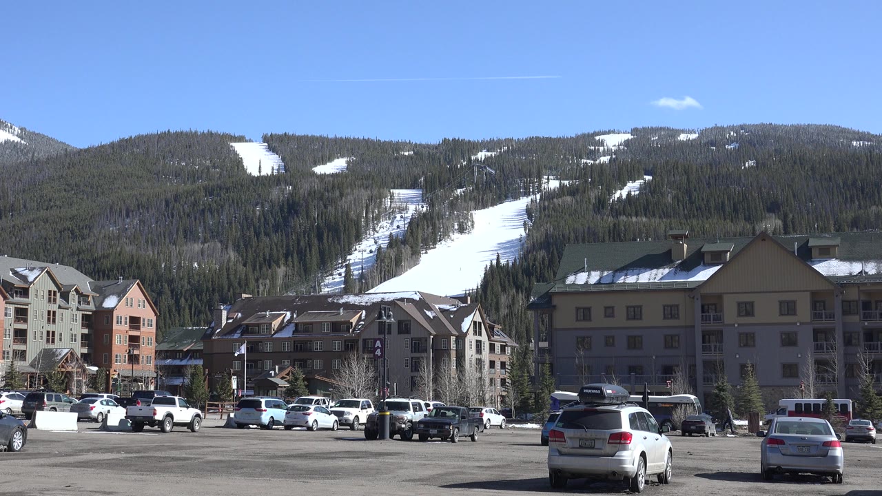 Parking lot, lodge buildings, forested ski hill, and blue sky.