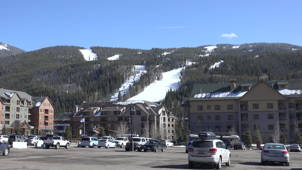 Opening frame of a Keystone, Colorado ski resort parking lot with parked cars, lodge-style buildings, forested slopes, white ski runs, clear blue sky, and no visible people.