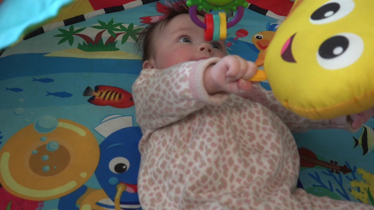 Infant reaching toward a yellow hanging toy on a colorful play mat.