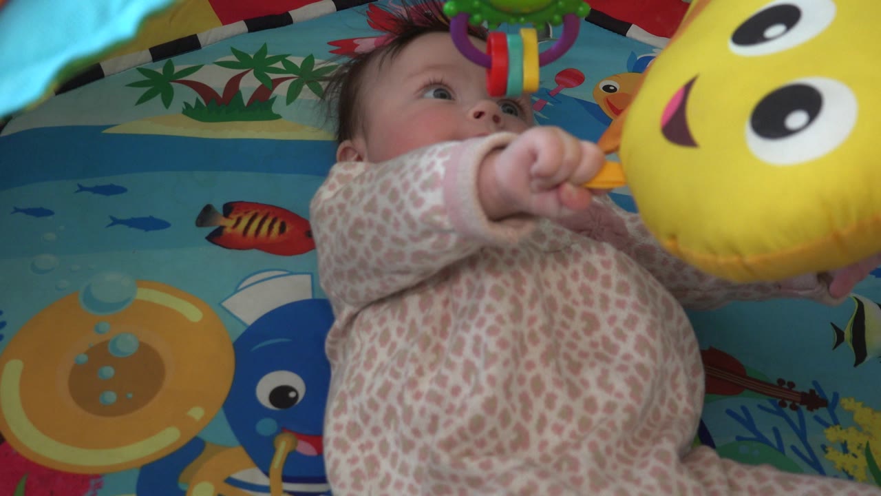 The infant looks upward from the play mat while one hand grips near the yellow toy, with ocean-themed fish graphics filling the background.