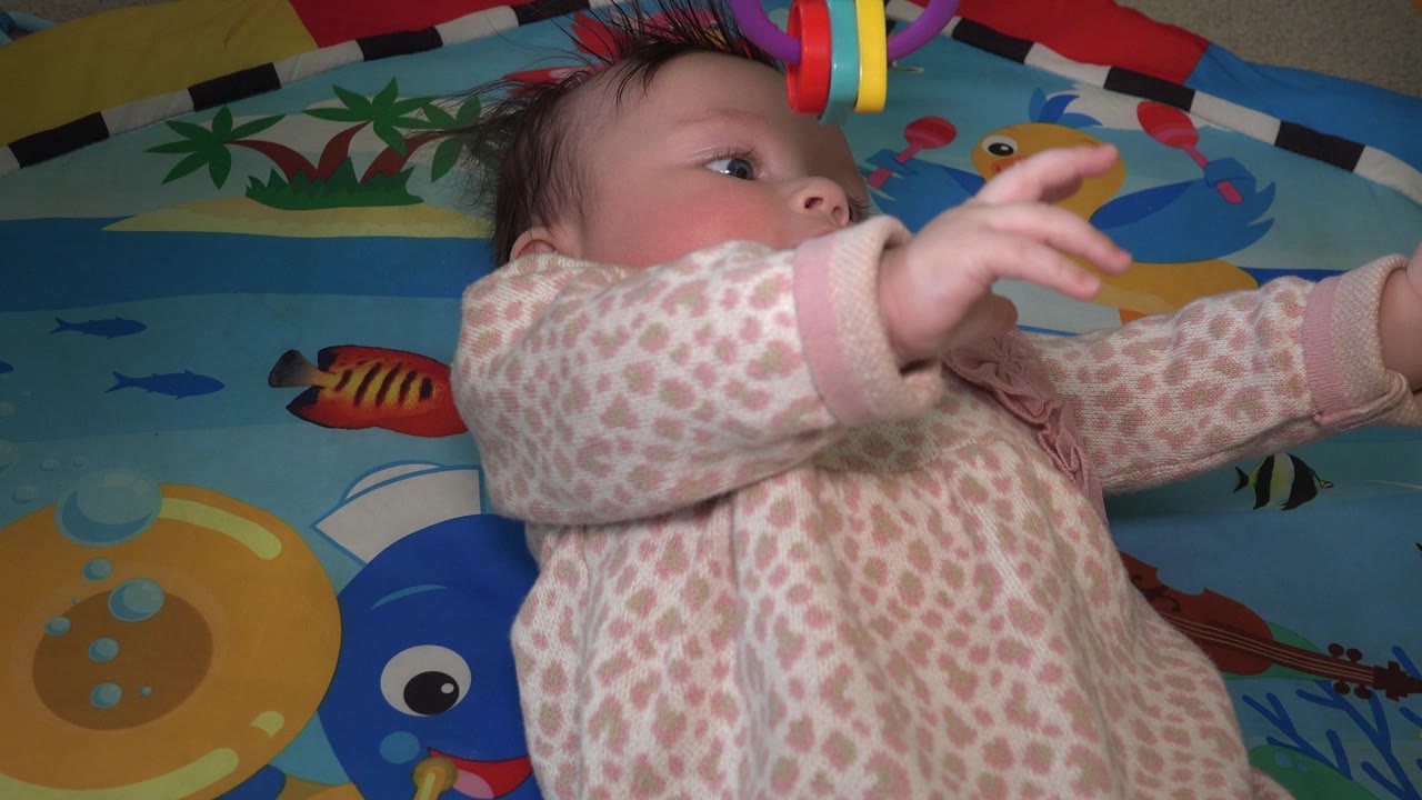 The infant remains on her back in Westminster, Colorado, with her arms lifted toward red, yellow, and purple toy rings above the play mat.