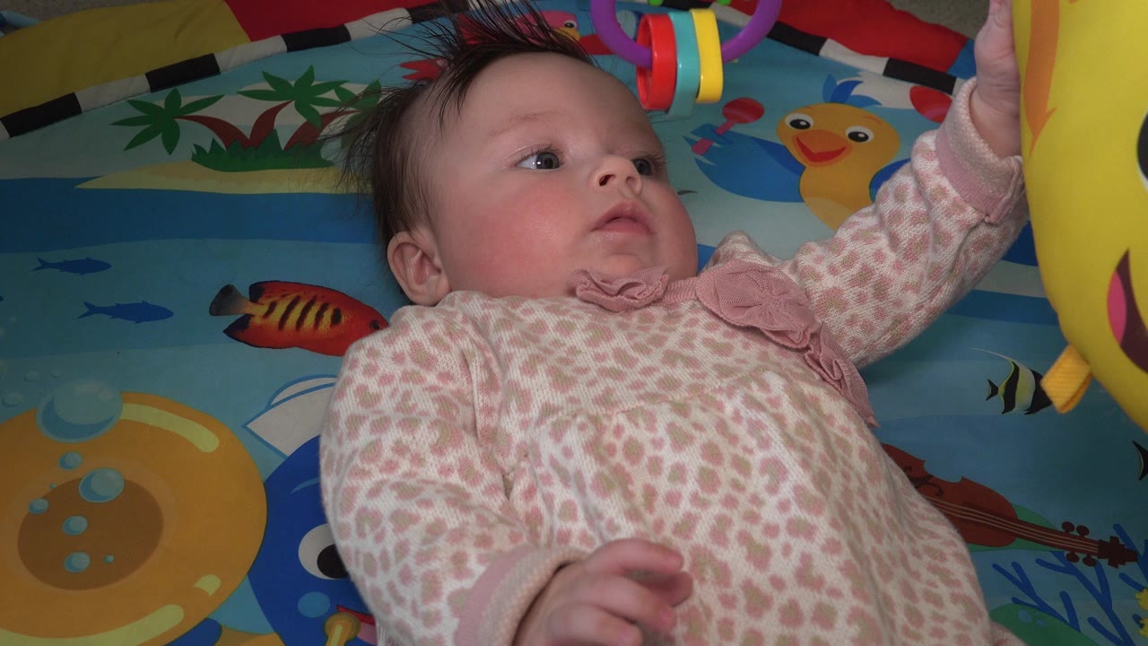 Baby girl lying on a colorful play mat reaching toward a yellow hanging toy.