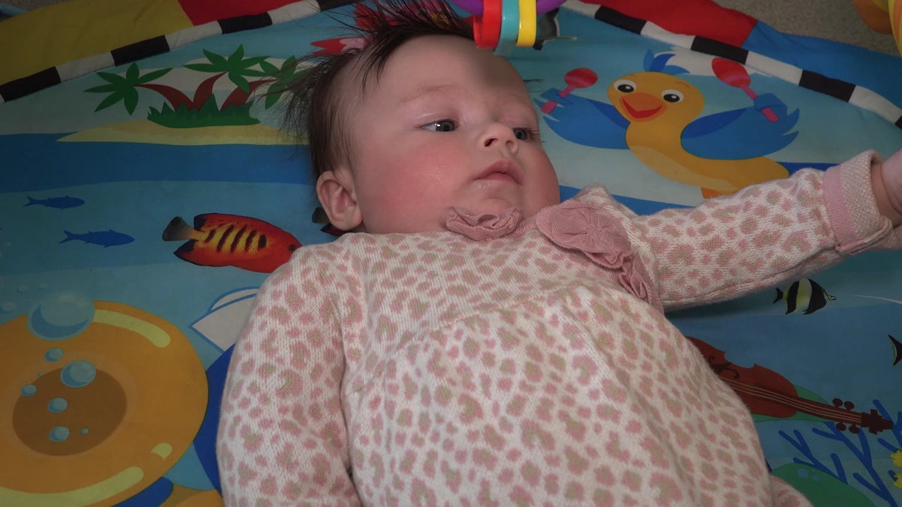 Young child lying on a blue play mat under colorful hanging rings.
