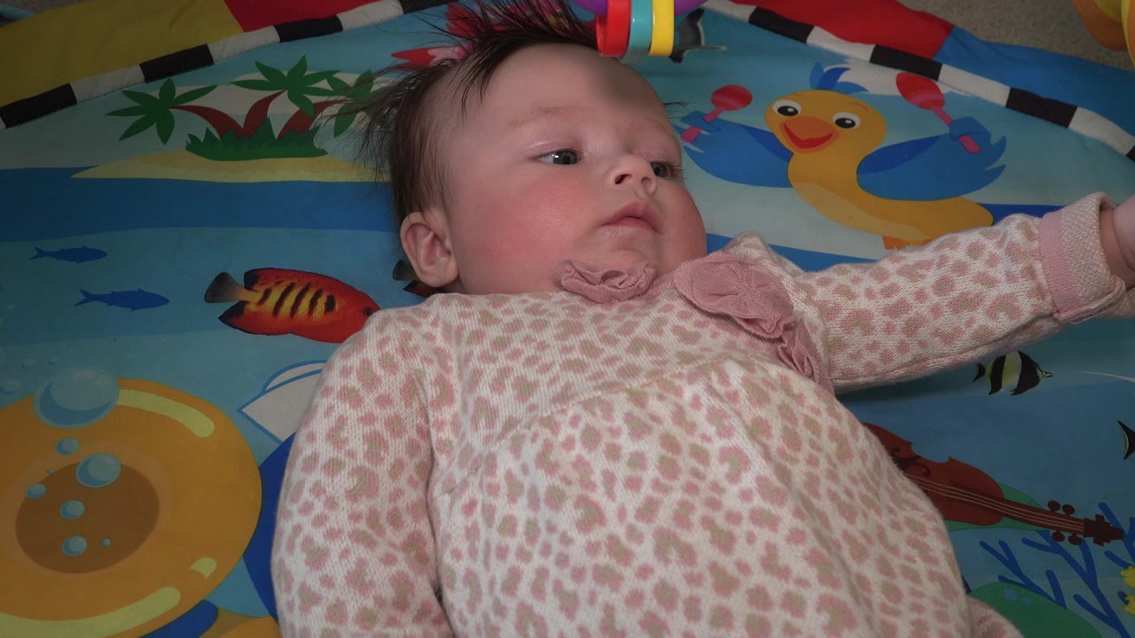 The child remains on the blue play mat in Westminster, Colorado, looking upward while the colorful ring toy hangs near the top edge of the frame.