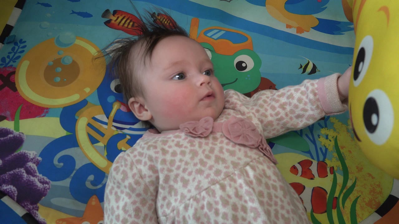 Baby in a pink and white sleeper lying on a colorful play mat near a yellow stuffed toy.
