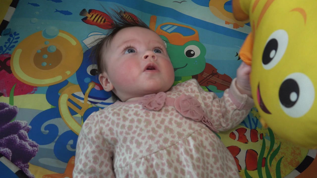 Baby girl lying on a colorful play mat beside a yellow stuffed fish.