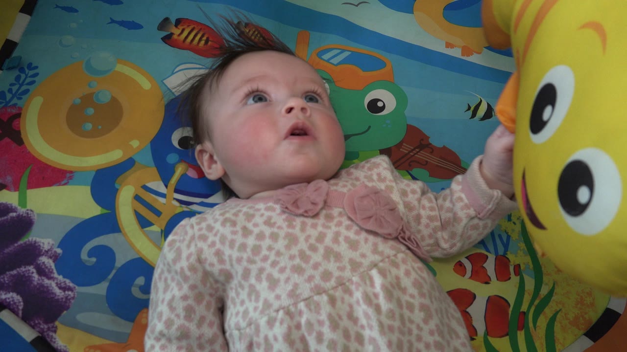 At 0:01 in Westminster, Colorado, the baby remains on her back, eyes lifted, with one hand near the oversized yellow stuffed fish and cartoon sea graphics around her.