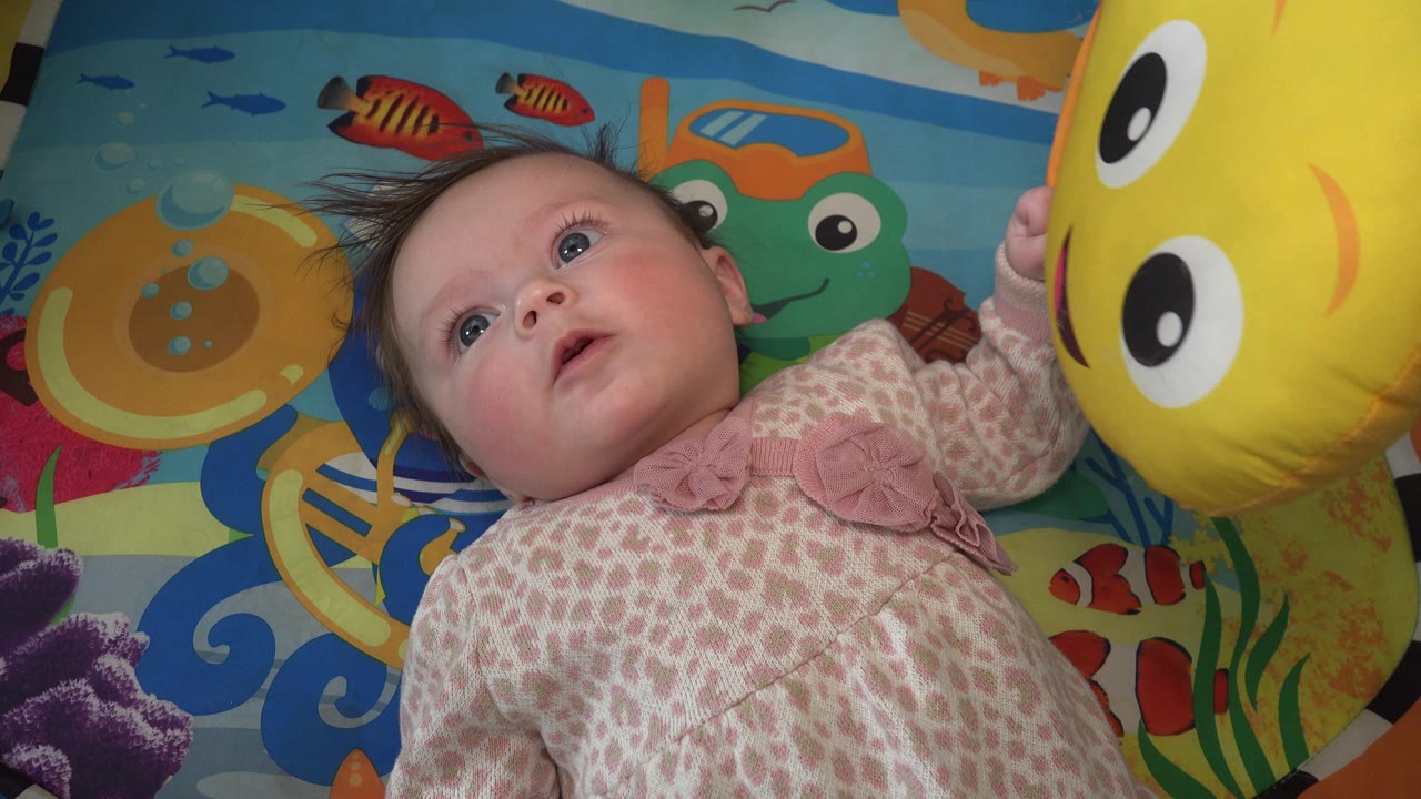 A baby in Westminster, Colorado looks forward with one hand raised toward the large yellow happy-face balloon entering from the right.