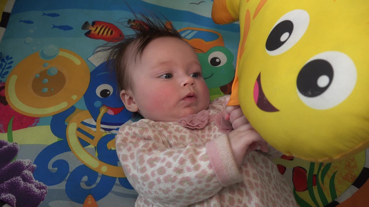 The baby remains on the patterned play mat in Westminster, Colorado, with one hand near the yellow toy and eyes turned toward it.