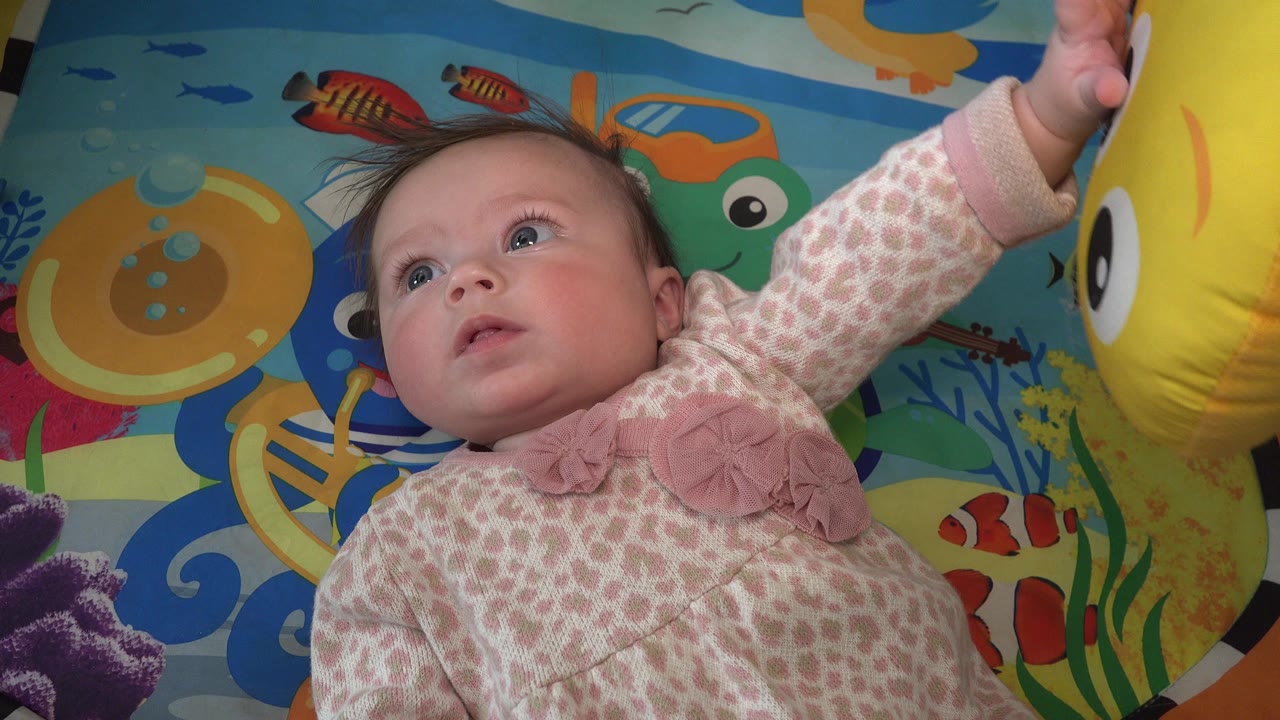 In Westminster, Colorado, the baby girl keeps her eyes lifted and extends one arm upward beside a large yellow toy over the colorful play mat.