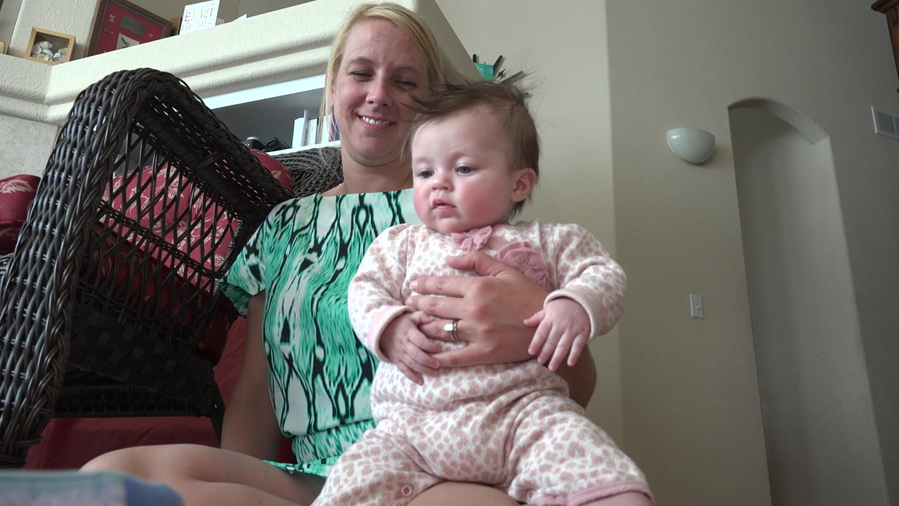 Woman holding a baby on a red cushioned wicker chair indoors.