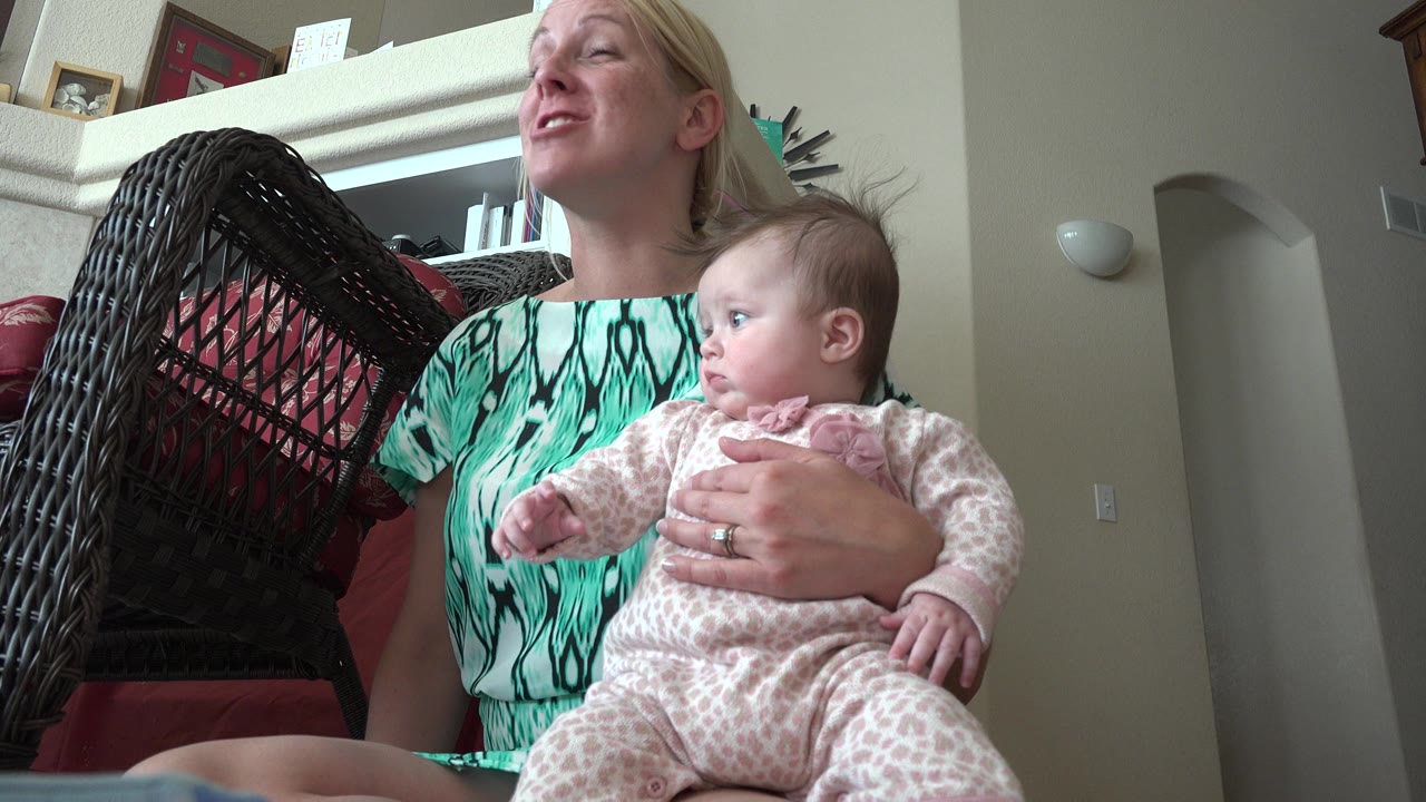Mother holding a baby indoors beside a wicker chair.