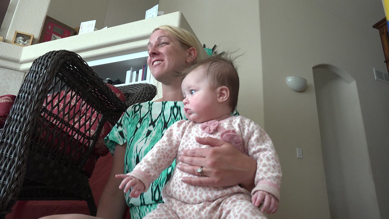 The woman continues holding the baby in the Westminster, Colorado home while the baby looks off to the right and the wicker chair fills the left side of frame.