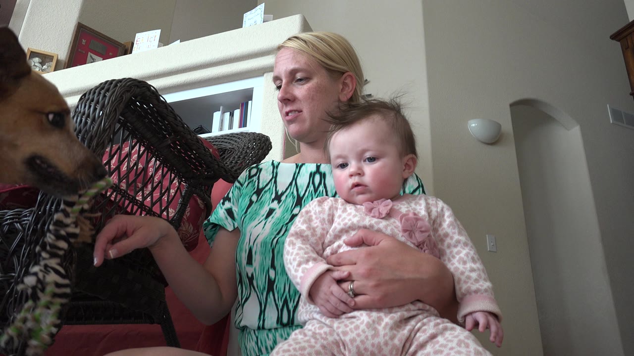 Woman holding a baby as a dog appears at the left edge indoors.