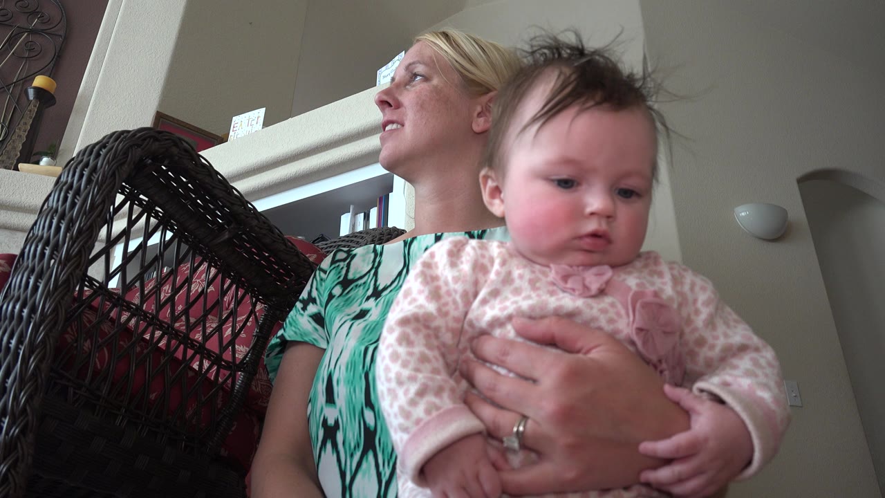 Baby held by an adult woman beside a wicker chair indoors.