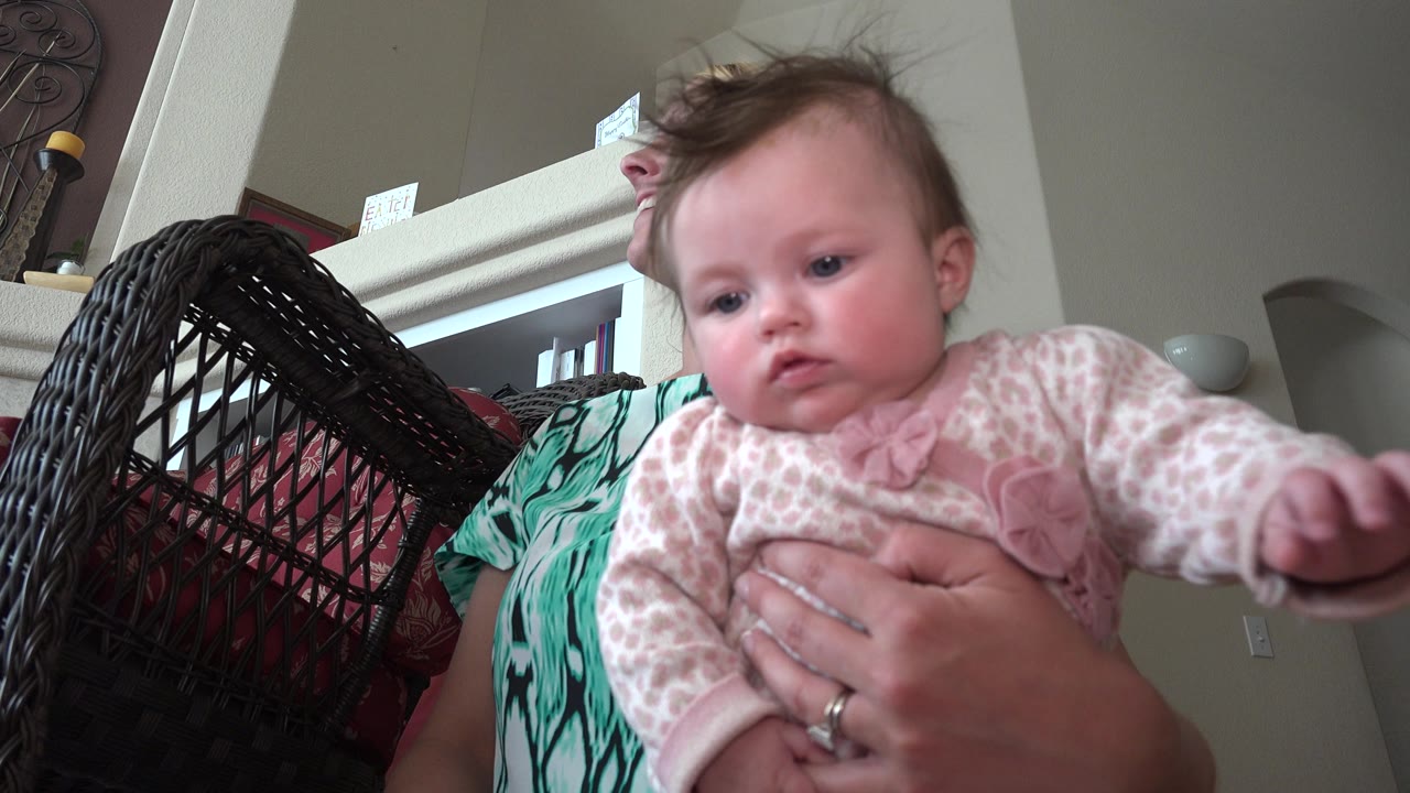 Baby in pink clothing held close to the camera by a blonde woman indoors.