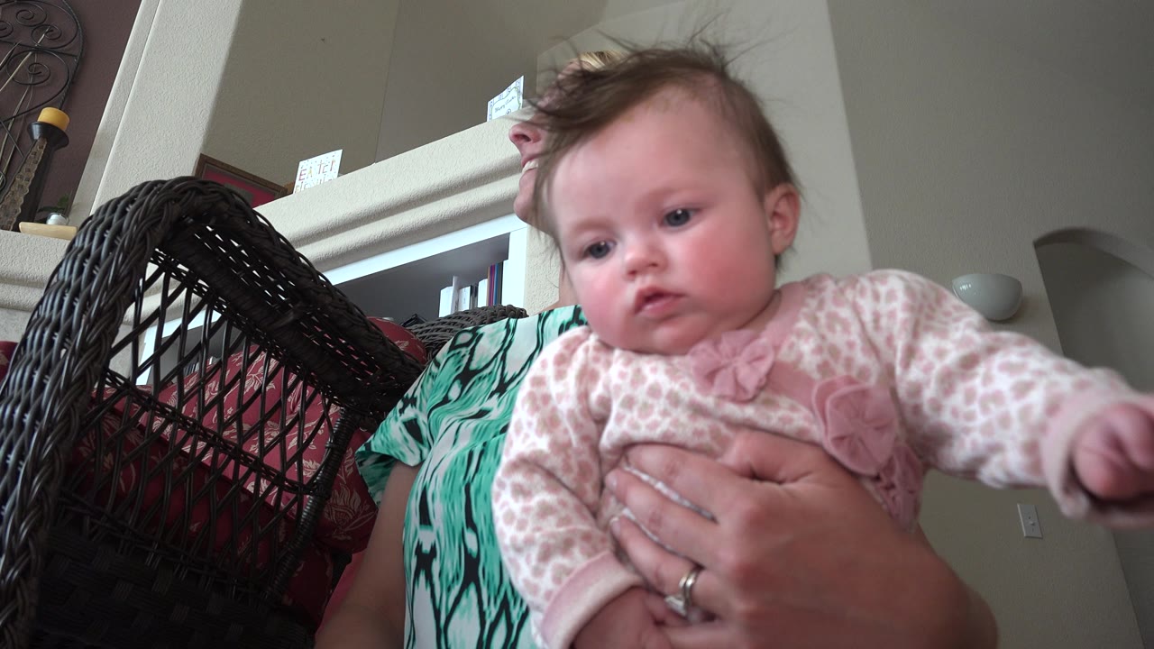 At 0:01 in Westminster, Colorado, the baby extends an arm toward the camera while the blonde woman supports the child from behind.