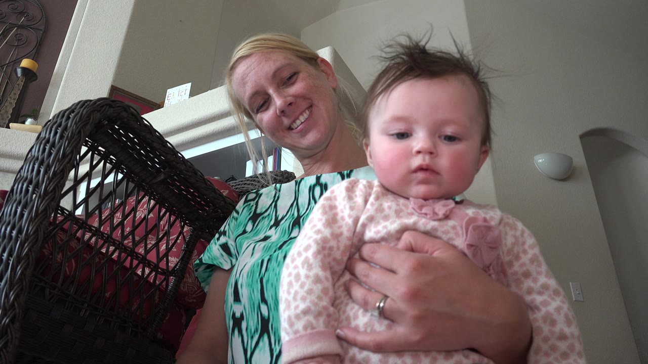 Woman holding a baby indoors beside a dark wicker chair.