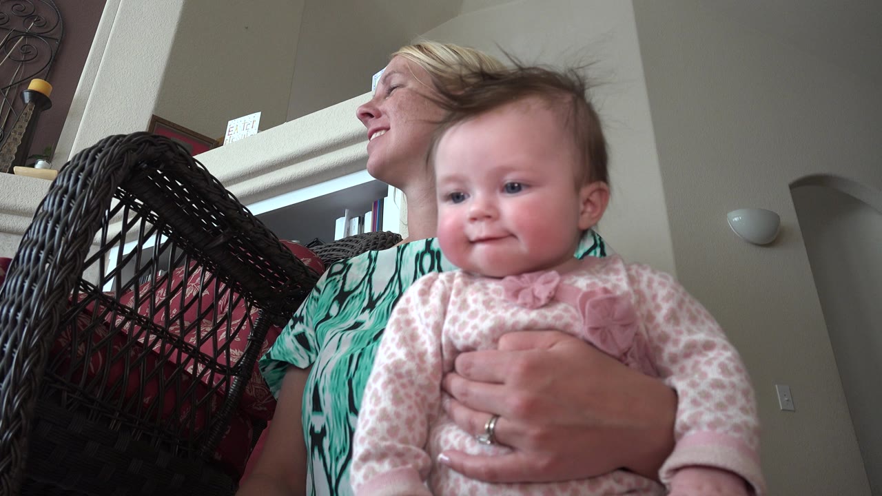Woman holding a baby indoors beside a wicker chair.