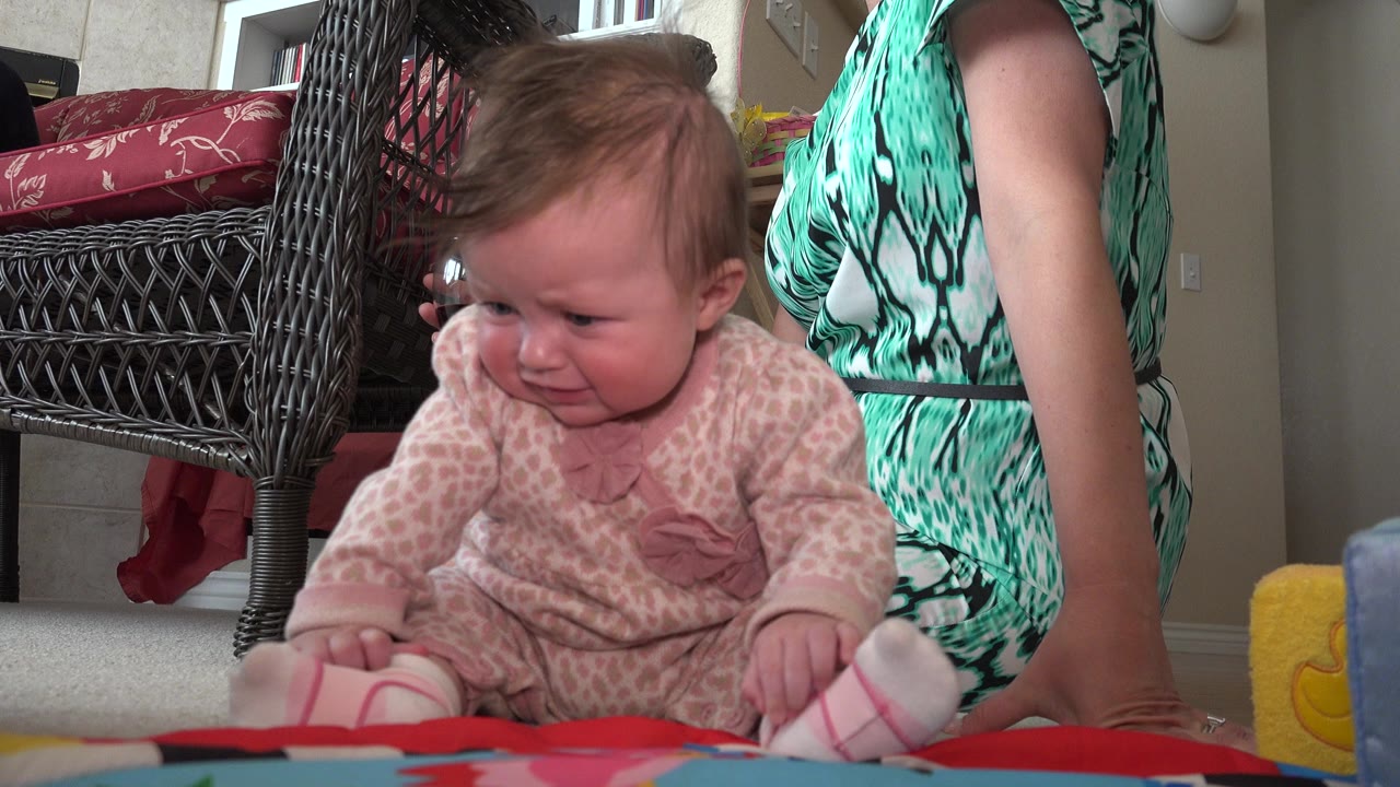 Baby sitting on a floor mat beside a seated woman indoors.