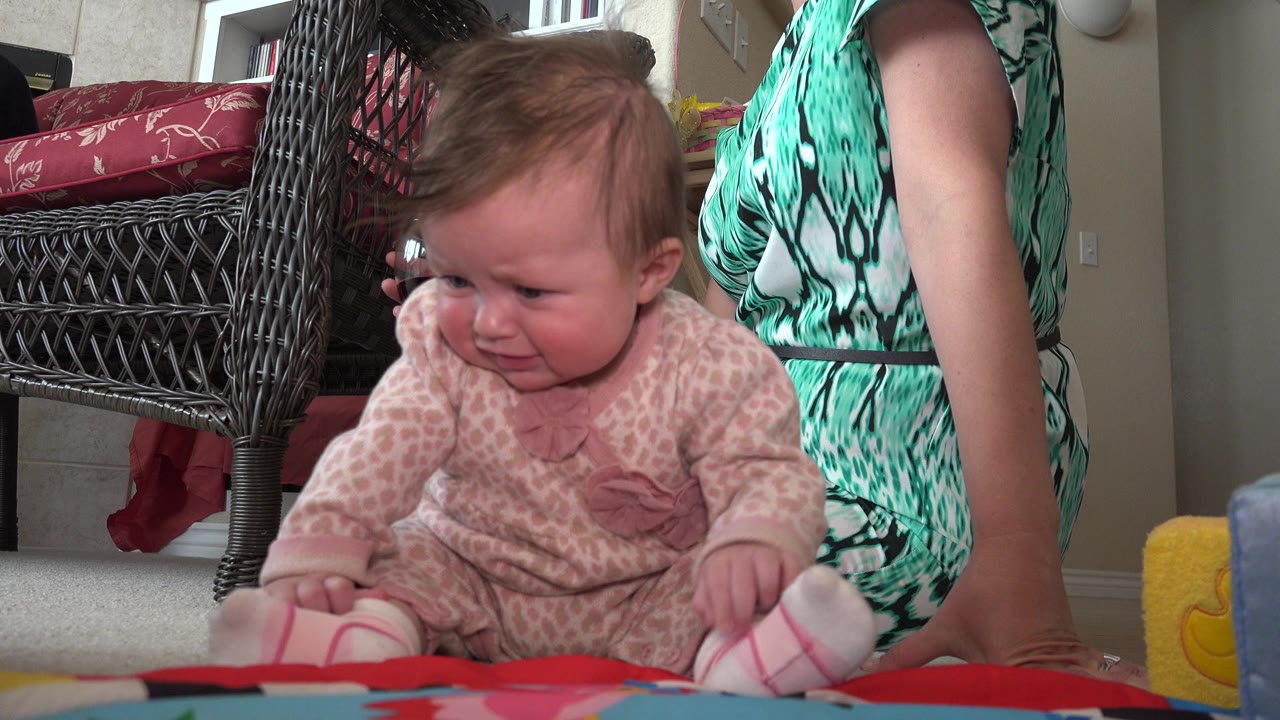 The baby leans slightly forward on the mat in the Westminster home interior, with the woman's torso and arm visible at the right edge and wicker furniture behind.