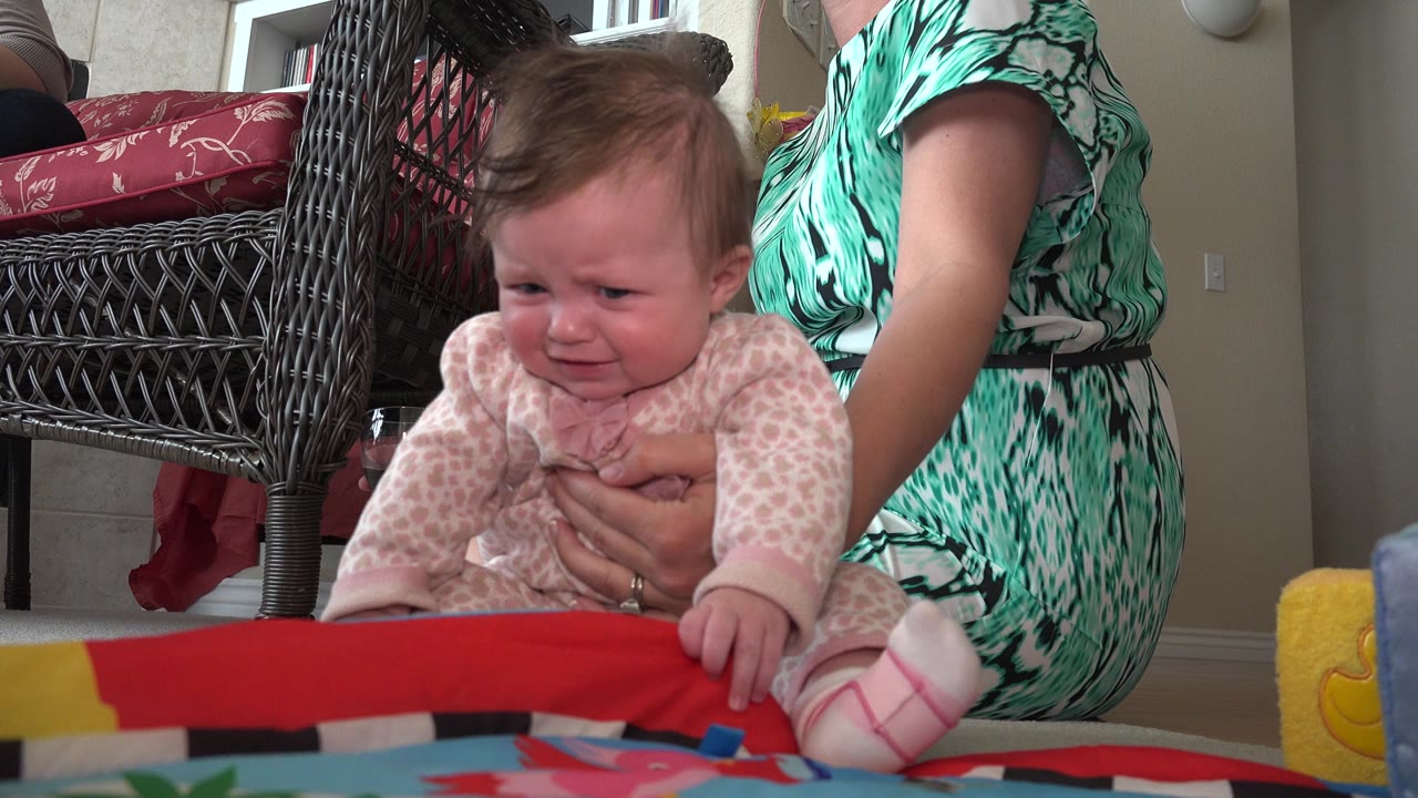 Infant leaning forward on a colorful mat while an adult supports her.