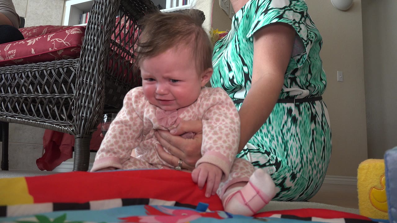In Westminster, Colorado, the infant's face is tearful as an adult hand supports her torso beside the wicker chair and patterned play mat.