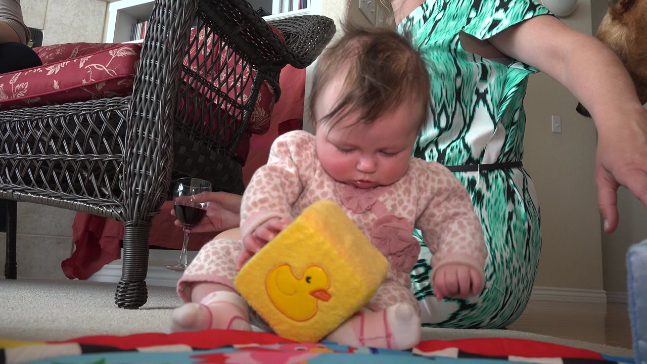 Baby holding a yellow duck cube beside an adult and a wine glass indoors.