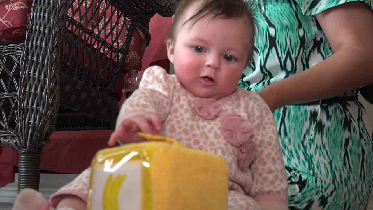 At 0:00, a baby in pink patterned clothes faces the camera in Westminster, Colorado, with one hand extended toward a bright yellow object in the foreground and an adult partly visible behind.