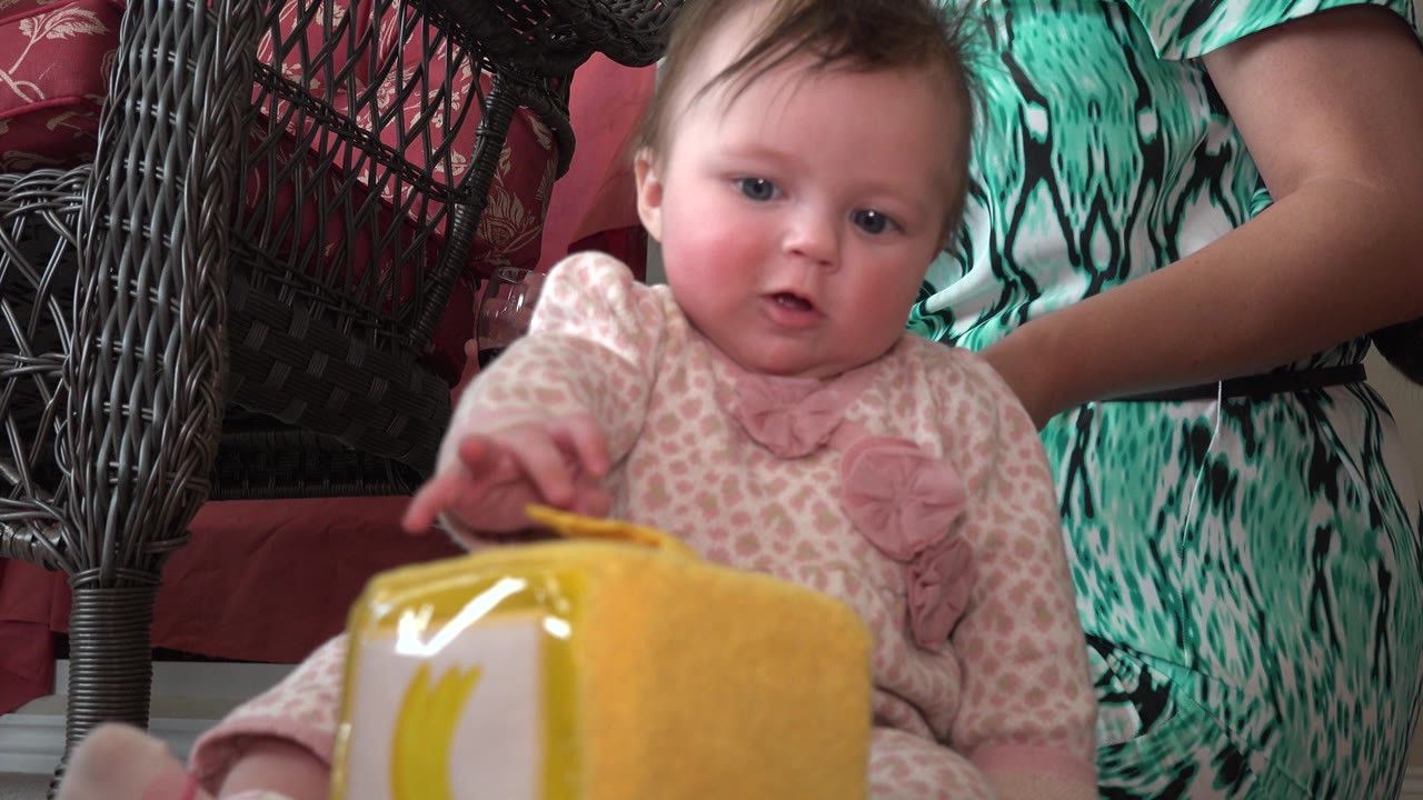 At 0:01, the baby remains centered and close to the lens, looking forward while a yellow object sits at the bottom of frame and an adult in green patterned clothing supports from behind.