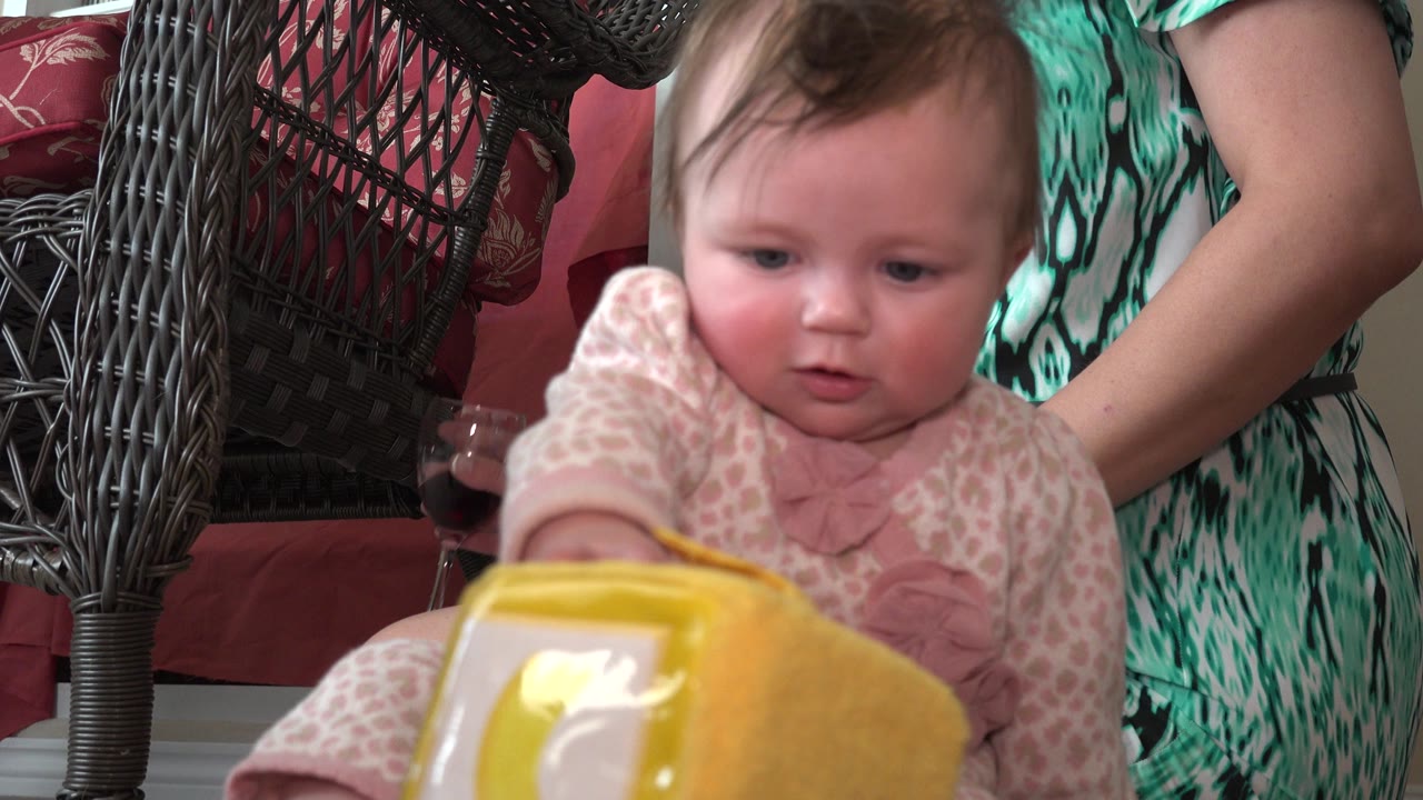 At 0:00 in Westminster, Colorado, a baby sits close to a yellow toy block while an adult's arms and green patterned dress appear behind them.
