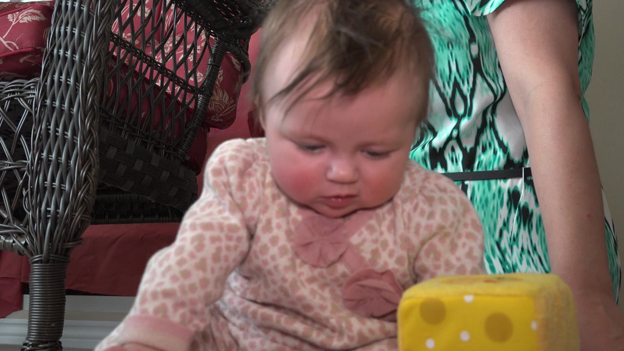 At 0:00, a light-skinned infant in a pale patterned onesie sits in a Westminster, Colorado home interior near a yellow toy block, with wicker furniture and an adult's arm partly visible.