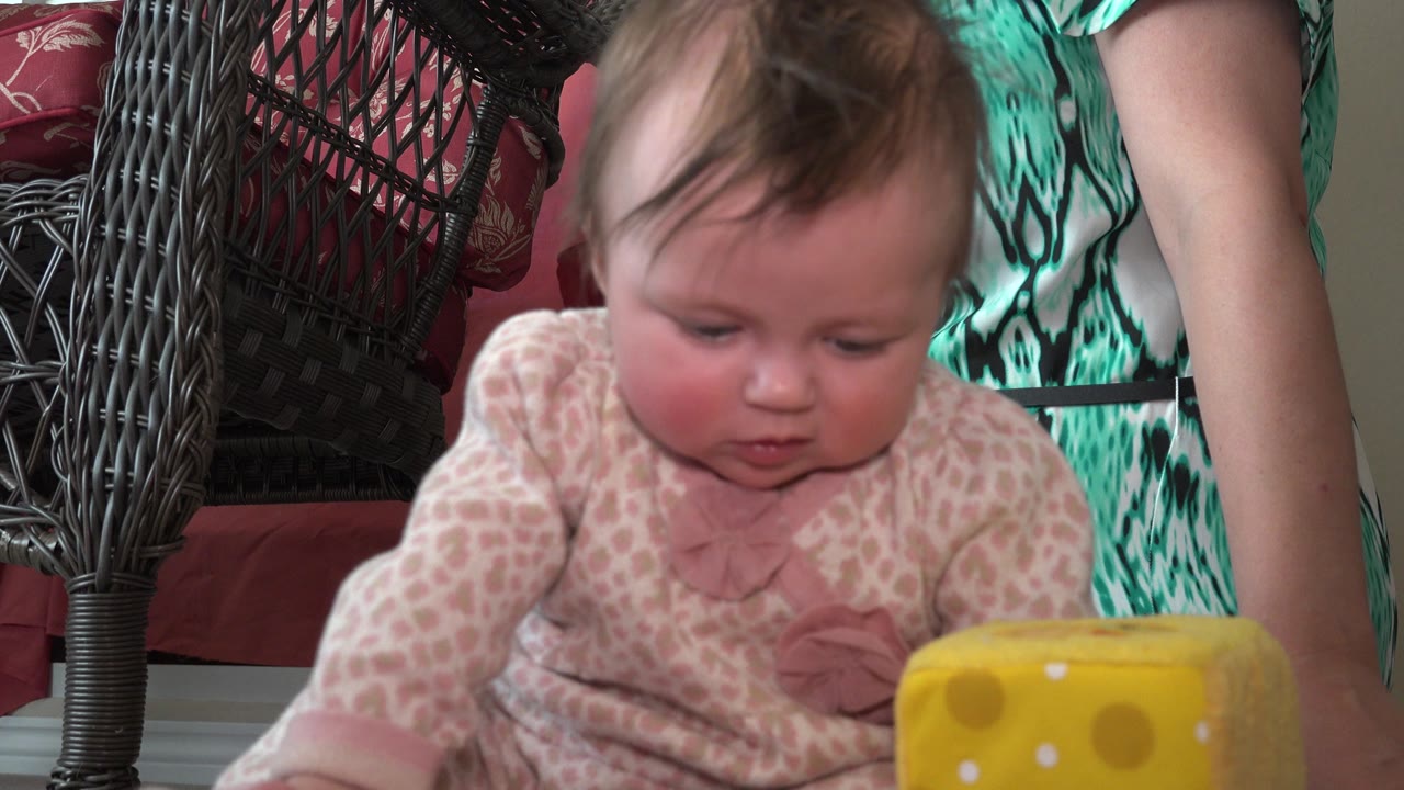 At 0:01, the infant remains centered and leans slightly toward the yellow toy block while an adult's torso and arm stay at the right edge of the Westminster home frame.