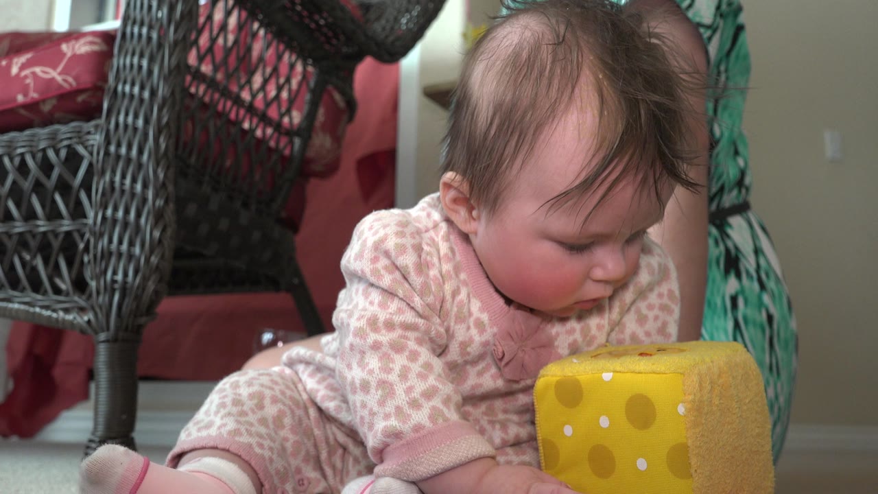Baby in pink and white sleeper beside a yellow plush dice toy indoors.