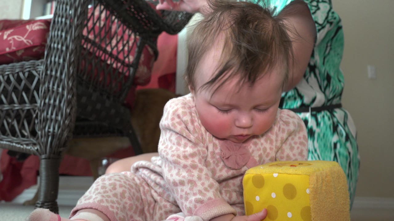 The toddler remains low on the carpet in the Westminster, Colorado interior, leaning toward the yellow toy cube as an adult's patterned clothing appears in the background.