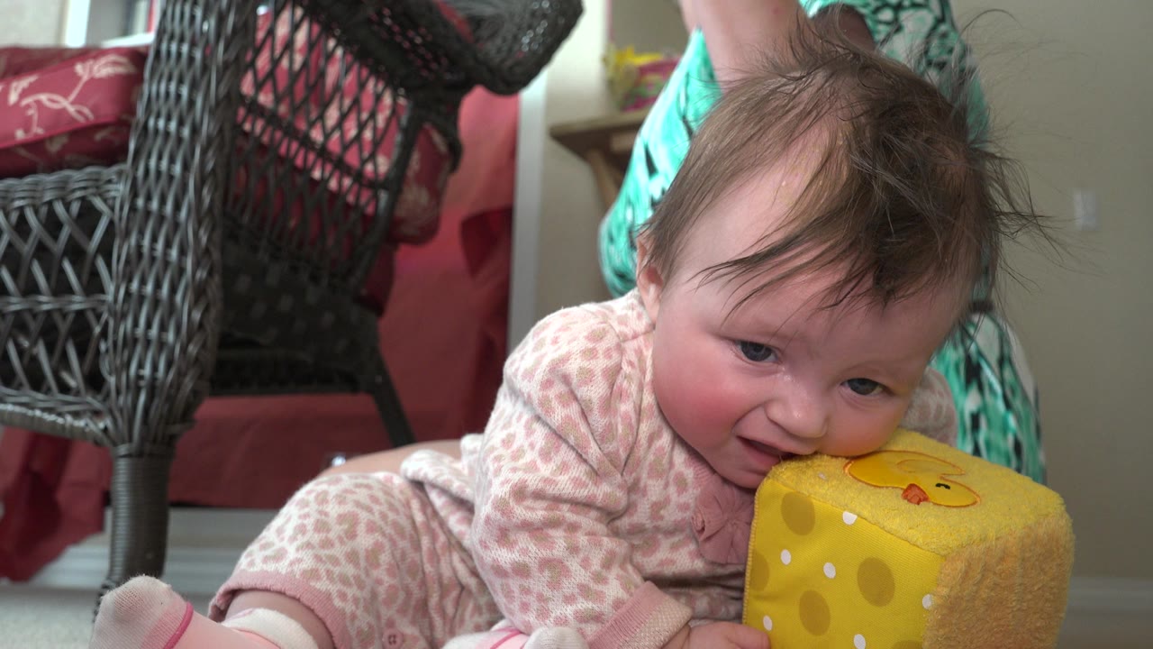 The baby brings their mouth to the yellow plush cube while seated low on the carpet, with a wicker chair and partial adult figure behind them in Westminster, Colorado.