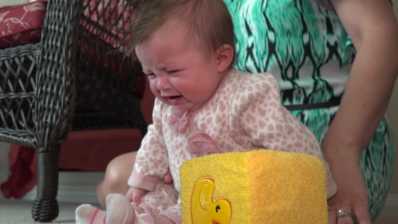 Crying baby seated beside a yellow soft block with an adult behind.