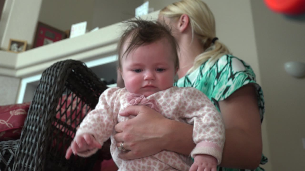 Baby in pink outfit held by woman indoors.