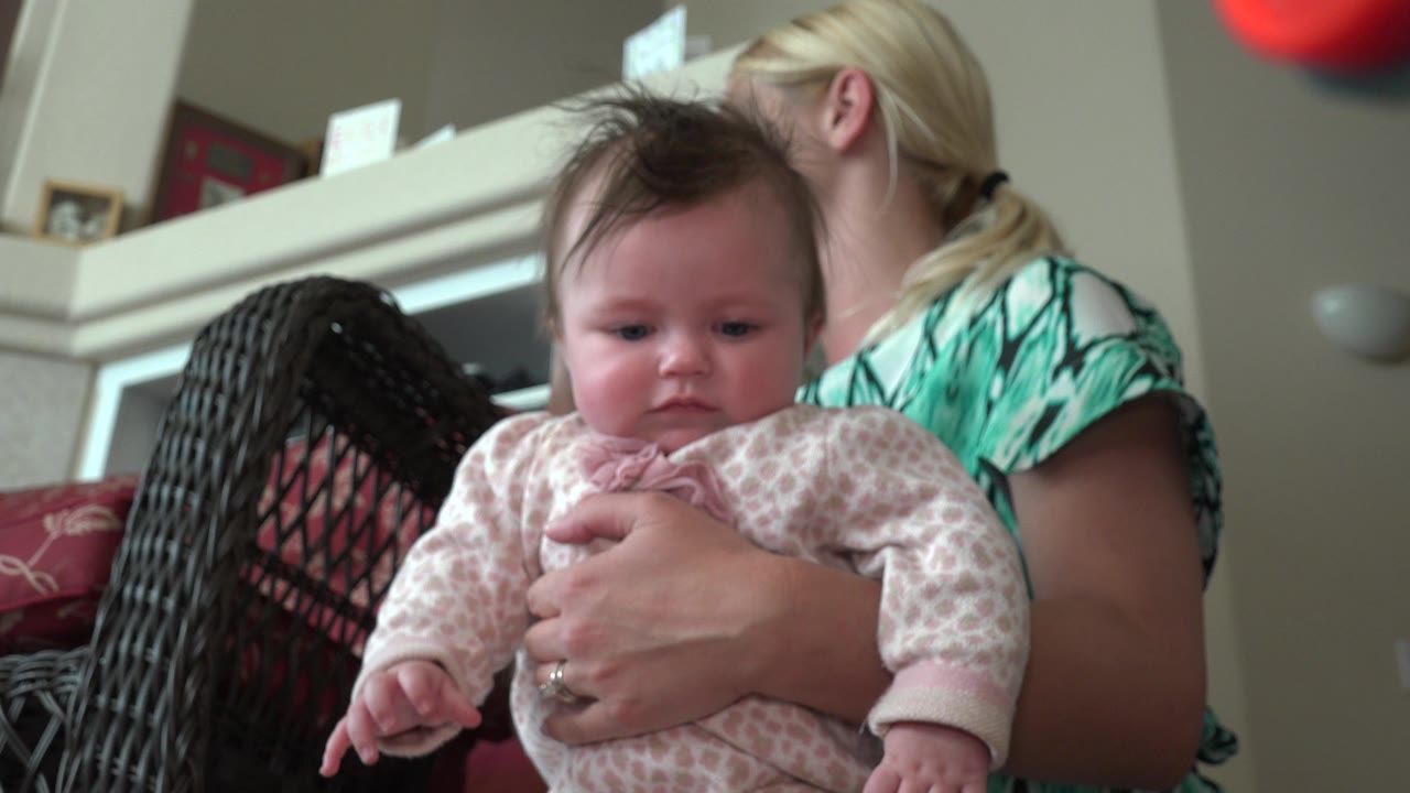 At 0:01 in Westminster, Colorado, the infant remains centered in the foreground, held around the torso, with the woman turned sideways behind a dark wicker chair.