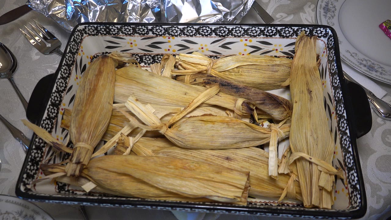 At 0:00 in Westminster, Colorado, a patterned rectangular casserole dish filled with corn husk-wrapped food sits among plates, forks, foil, and a pale tablecloth, with no visible people.