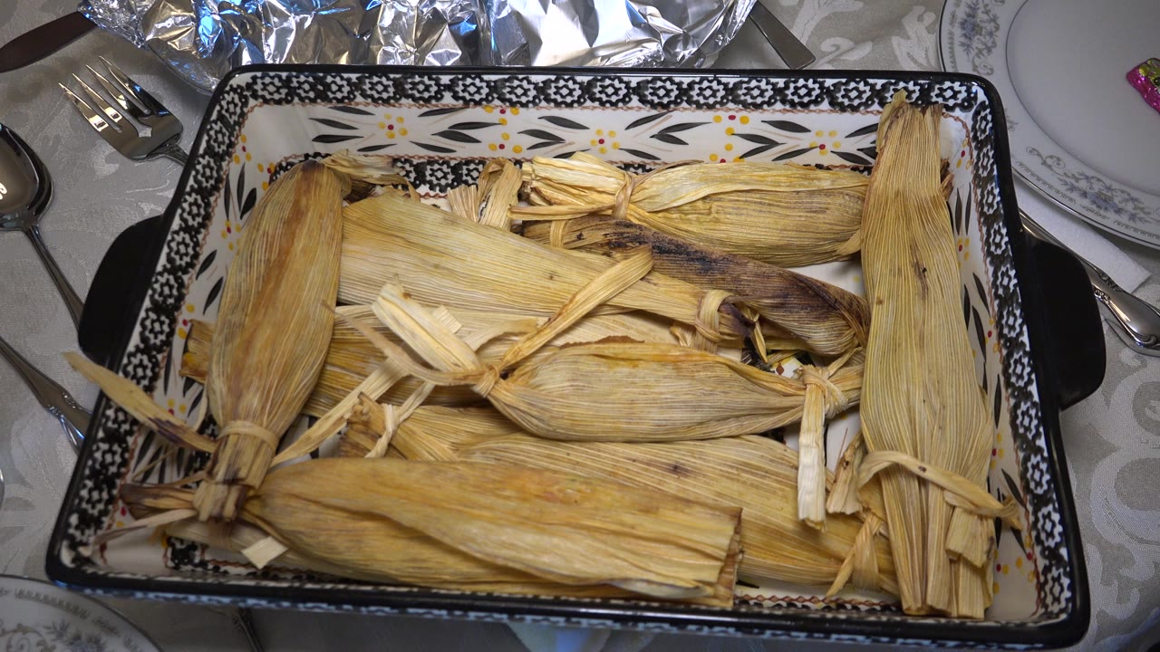 At 0:01, the serving dish remains centered with tan corn husks layered across it, black-and-white rim patterns visible, and no visible people around the Westminster dinner table.