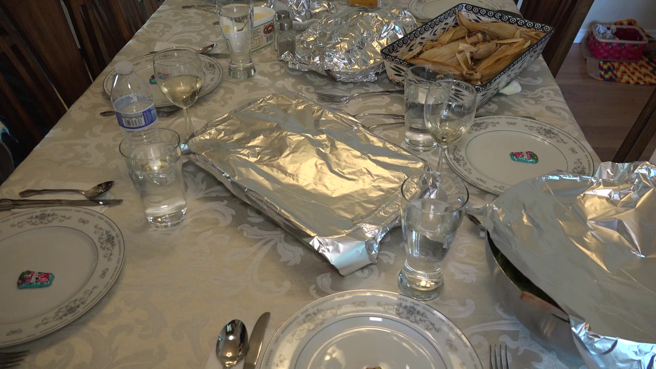 At 0:01, the view remains angled over the Westminster dining table, with reflective foil trays centered between plates, water glasses, wine glasses, spoons, and a basket of bread or pastries, with no visible people.