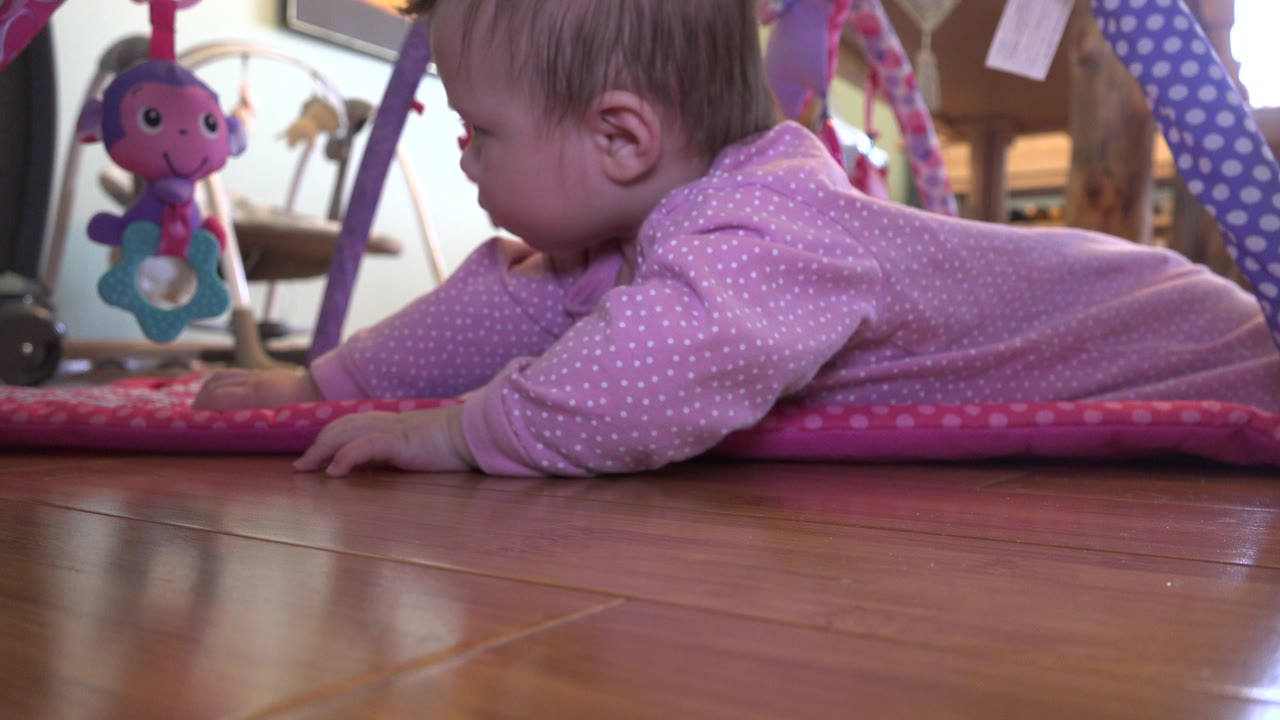 At 0:00 in Westminster, Colorado, a baby in a pink polka-dot sleeper lies on a pink play mat at floor level with a hanging toy beside the face.