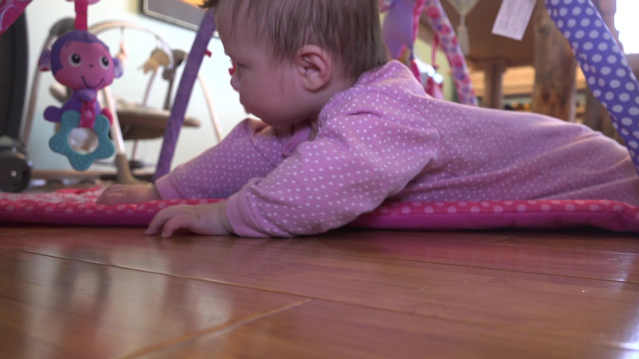 At 0:01 in Westminster, Colorado, the baby remains stretched across the pink mat in side profile while the hanging toy stays near the left side of the frame.