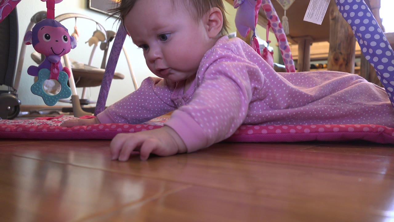 At 0:00, a baby in a pink jumpsuit lies on a padded play mat in Westminster, Colorado, reaching toward the wood floor with a hanging toy beside the face.