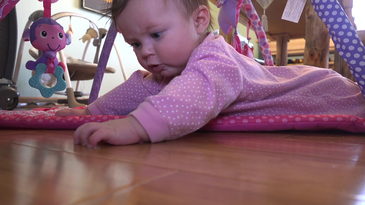 At 0:02, the baby in Westminster, Colorado looks down toward the mat while lying on the stomach, with pink fabric, toy straps, and the wood floor filling the frame.