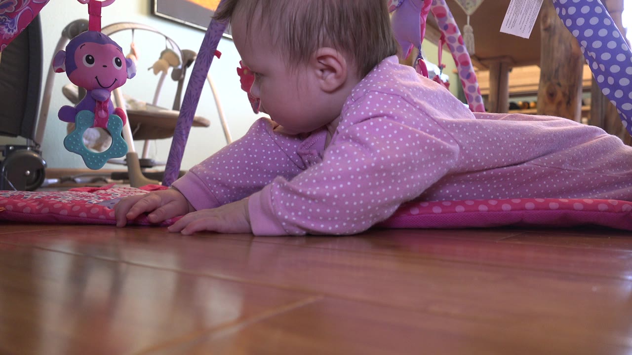 The baby remains low on her stomach on the pink mat in Westminster, Colorado, with both hands forward and a smiling plush toy hanging in front of her.