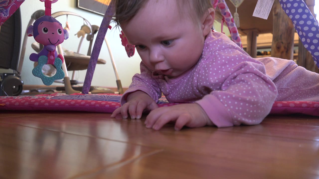 Baby in a pink sleeper lying on a wooden floor near a play mat.