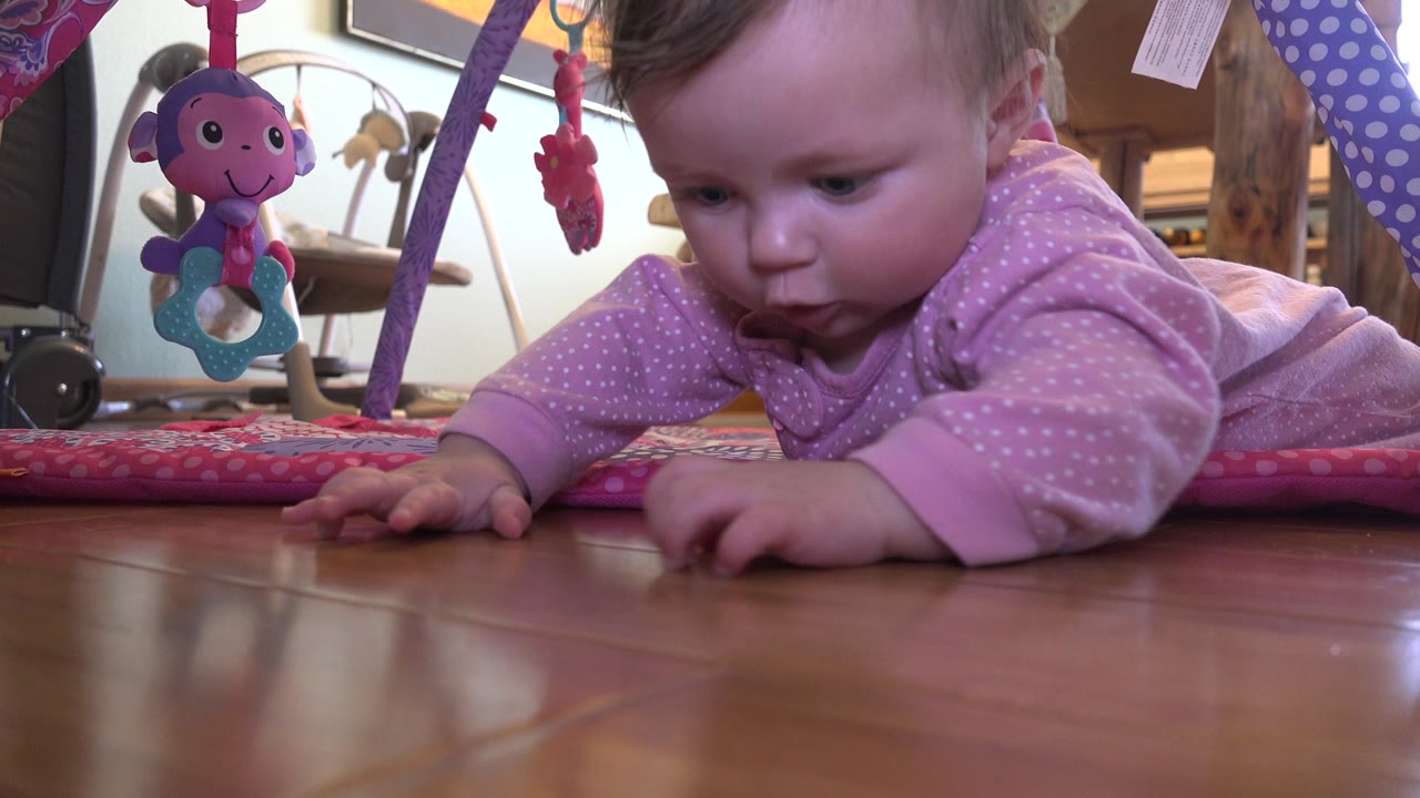 Baby in a pink sleeper lying on a hardwood floor near hanging toys.
